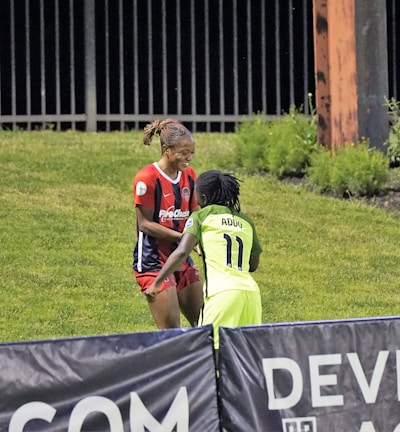 Two individuals are interacting on a grassy field. One is wearing a red and blue sports outfit with 'ProChain' visible, and the other is in a bright yellow uniform with the name 'Addo' and the number 11. They appear to be playing a sport, possibly soccer, as they are near a marked boundary. A fence and some vegetation are visible in the background.