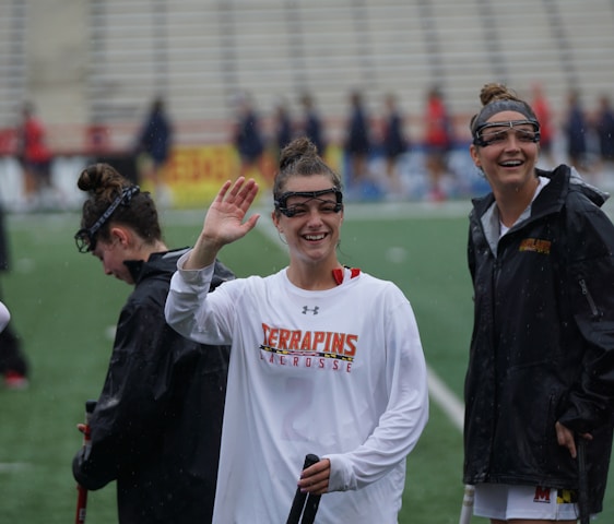 Several athletes in sports attire are on an outdoor sports field, interacting and smiling. One individual in the foreground waves with a friendly expression, while another stands beside her, also smiling. They wear protective goggles and gear associated with lacrosse. The background features indistinct figures in dark clothing against a backdrop of stadium seating.