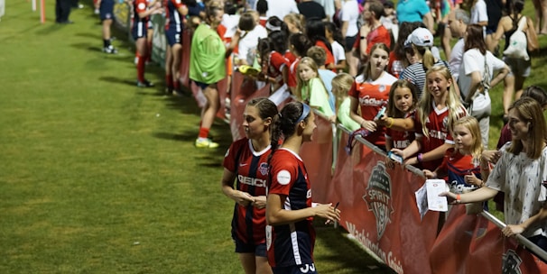 The image features a lively gathering of young soccer players and fans next to a field. Several female athletes in red and blue uniforms interact with an enthusiastic crowd standing behind a barrier. Some people in the crowd are holding objects, like pens and paper, possibly for autographs.