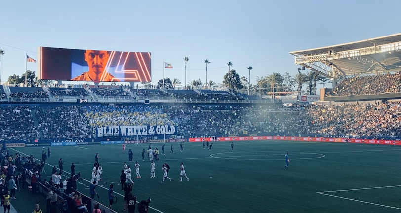 A large sports stadium filled with spectators. The field is occupied by soccer players, some walking and others scattered across the pitch. A massive screen displays a person, and flags fly on top of the stadium roof. The audience is dense, and the atmosphere seems lively with a visible banner showing 'BLUE WHITE & GOLD'.