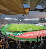 A large, brightly lit stadium filled with spectators. The field is green and well-maintained, with players actively engaged in a soccer match. The stands are crowded, and banners with writing are visible along the perimeter of the field. The atmosphere suggests excitement and energy.
