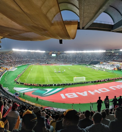 A large, brightly lit stadium filled with spectators. The field is green and well-maintained, with players actively engaged in a soccer match. The stands are crowded, and banners with writing are visible along the perimeter of the field. The atmosphere suggests excitement and energy.