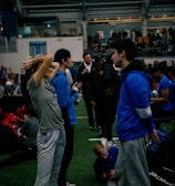 People engage in conversation and activities in an indoor sports facility. A woman in gray athletic wear stretches her arms above her head while talking to a man in a blue jacket. Other people are seen interacting in the background, some are seated on the ground, and the atmosphere is lively with many individuals gathered.