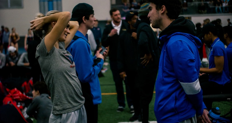 People engage in conversation and activities in an indoor sports facility. A woman in gray athletic wear stretches her arms above her head while talking to a man in a blue jacket. Other people are seen interacting in the background, some are seated on the ground, and the atmosphere is lively with many individuals gathered.