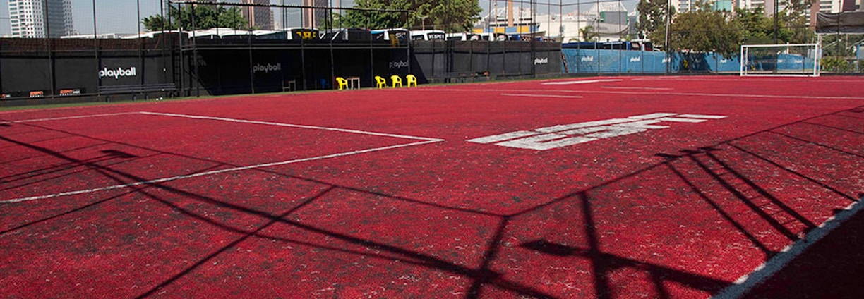 A sports court with a red playing surface and white lines, surrounded by a black mesh fence. In the background, tall buildings are visible, along with some trees and a few yellow chairs positioned near the fence.